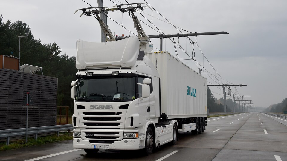 Un camion transporte un conteneur blanc sur une autoroute. Il est alimenté par des câbles électriques au-dessus de celui-ci.