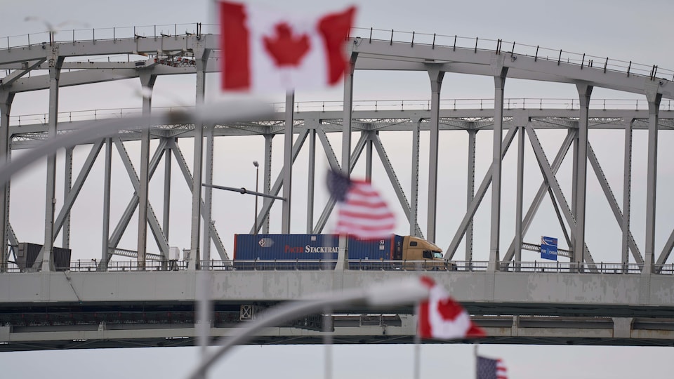 Les drapeaux du Canada et des États-Unis flottent au vent à la frontière entre les États-Unis et le Canada. On aperçoit, à l'arrière-plan, le Blue Water Bridge, qui relie Sarnia, en Ontario, à Port Huron, au Michigan.