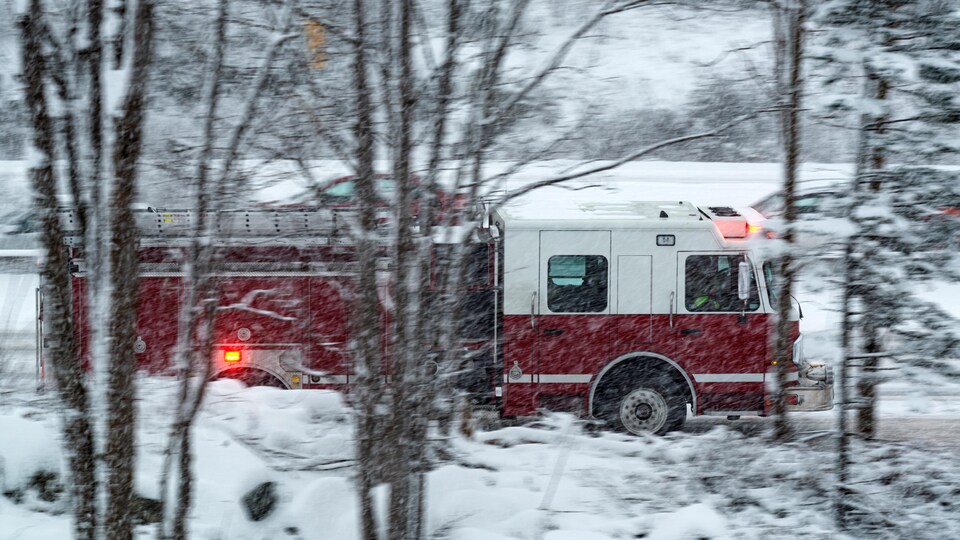 Un camion de pompiers en mouvement, ses gyrophares allumées, est photographié entre des branches d'arbres sur une route rurale enneigée.