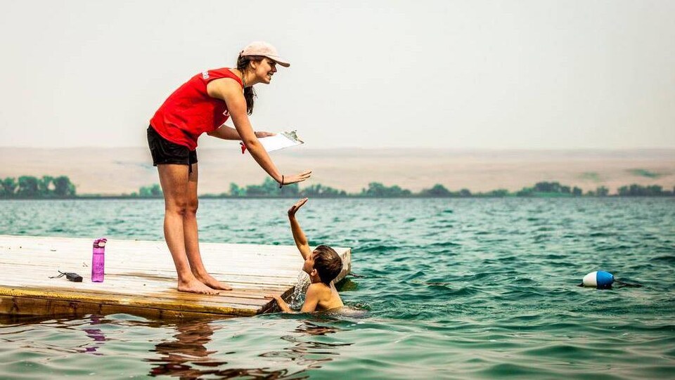 Un enfant se baigne dans un lac, il tape dans la main de sa monitrice qui est sur le quai.