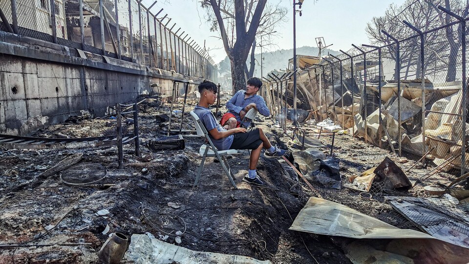 Deux hommes sont assis au milieu des décombres, entre deux clôtures entourant le camp.