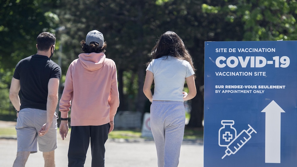 A man, a teenager and a teenage girl walk near a sign announcing a COVID-19 vaccination site.