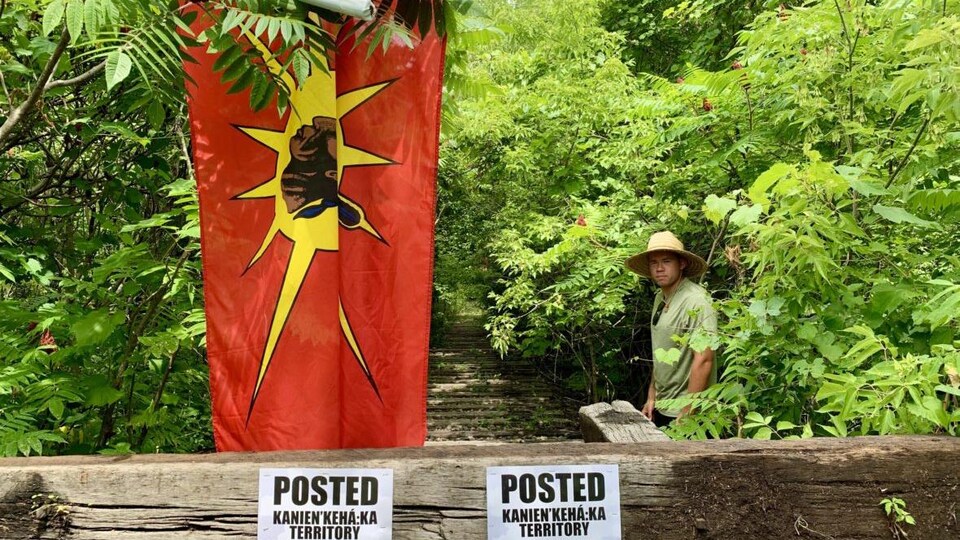 Un homme se tient près d'une route barricadée où est installé un grand drapeau mohawk.