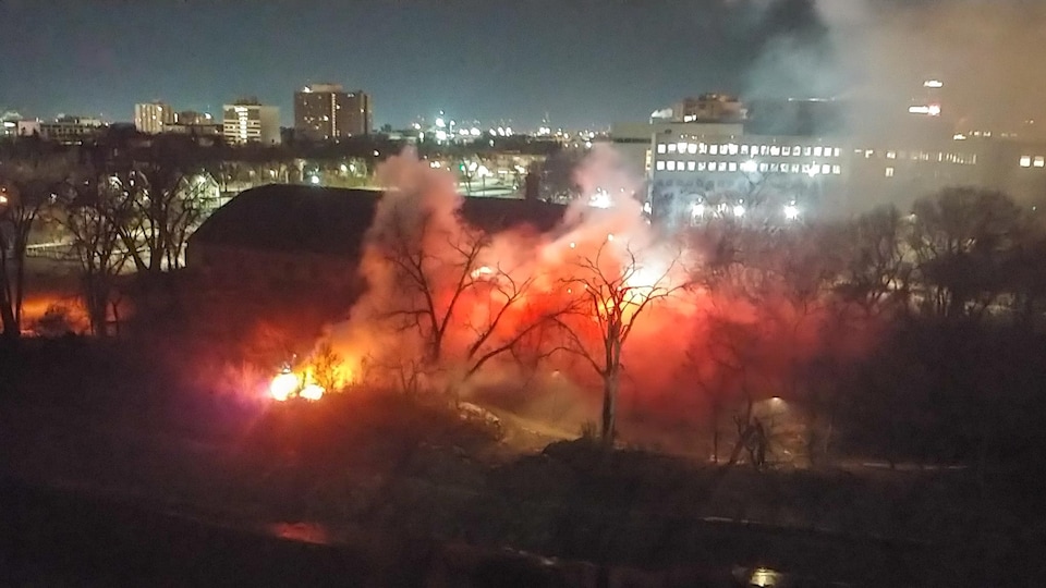 Des flammes et de la fumée entourent le bâtiment d’un club de curling près de la berge de la rivière, alors qu'il fait encore nuit.