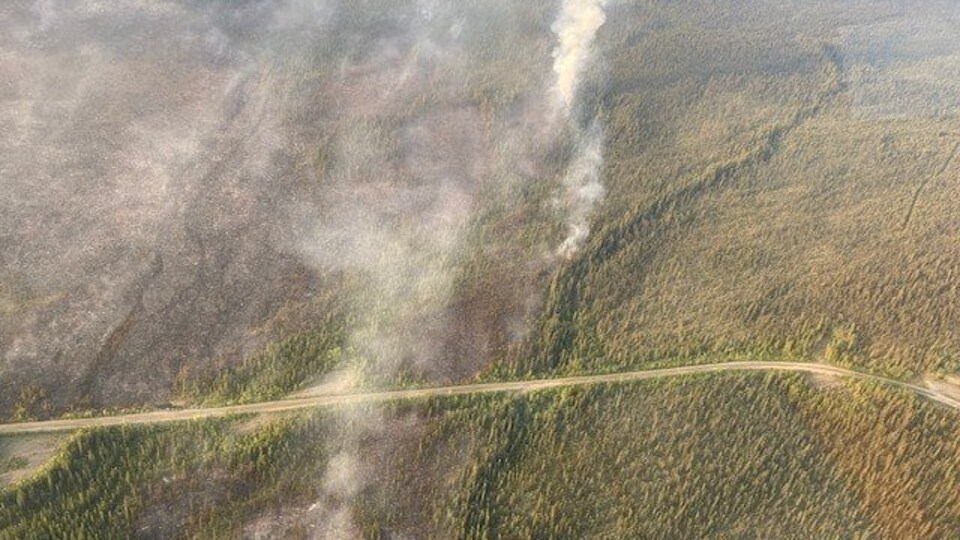 Un panache de fumée et une vue aérienne de la route Robert Campbell près de laquelle brûle un feu de forêt.