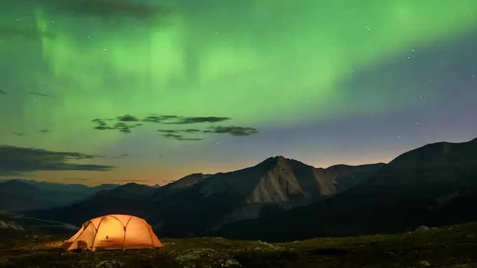 Stone Mountain Provincial Park in the northern Rockies. A tent is illuminated with mountains and the Northern Lights in the background.
