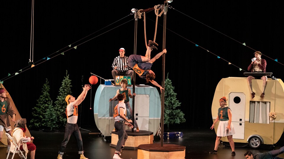 Des artistes de cirque performent dans un décor de camping avec des petites caravanes.