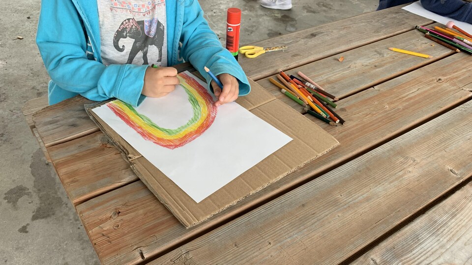 Une jeune fille dessine un arc-en-ciel coloré sur une table de pique-nique, où du matériel de bricolage est disposé. 