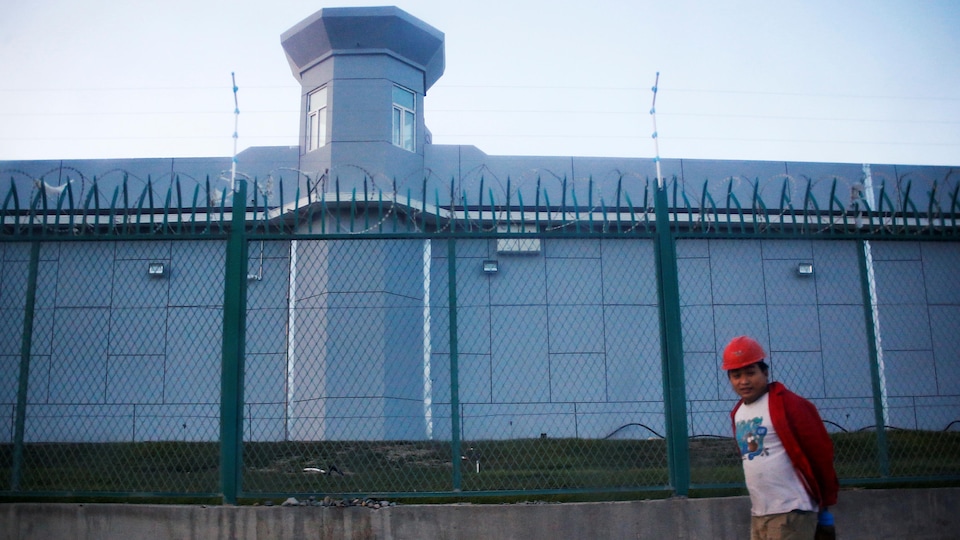 Un homme passe devant un établissement ceinturé par une haute clôture. Une tourelle de surveillance est aussi visible. 