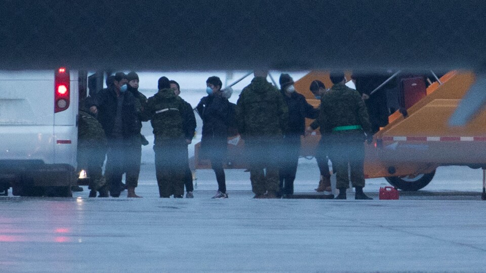 Des passagers masqués sont accueillis par des militaires sur le tarmac d'un aéroport. 