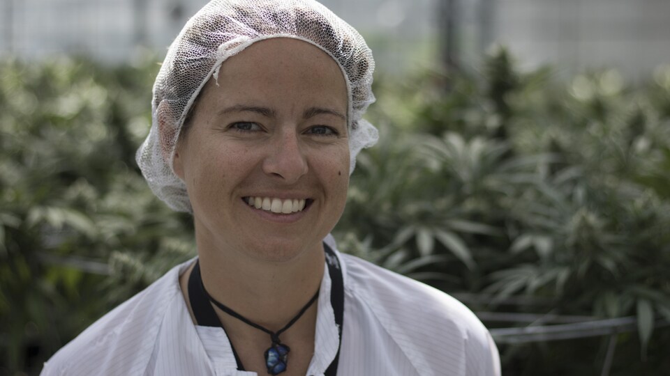 Une jeune femme portant un filet sur les cheveux et un sarrau blanc sourit à la caméra devant des plants de cannabis.