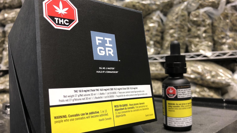 Une petite bouteille d'huile de cannabis à côté de sa boîte en carton, le tout devant un étalage de grands sacs de cannabis séché.