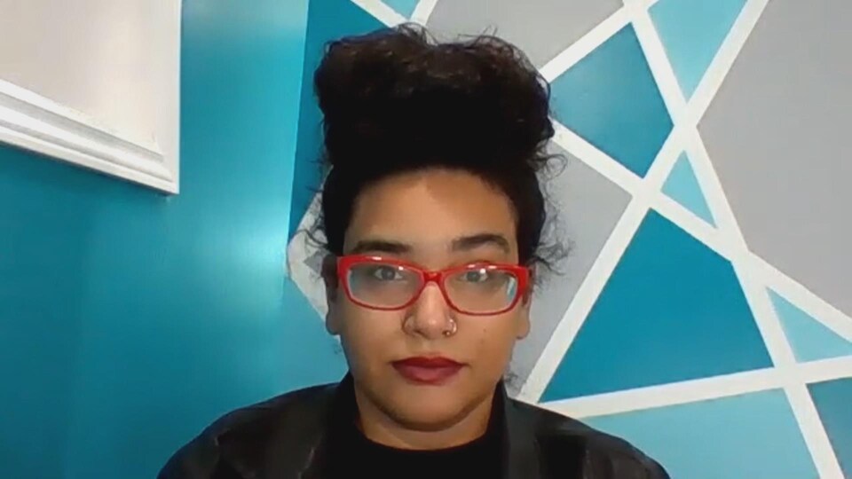 Une femme avec des lunettes, devant un mur bleu, en vidéoconférence.