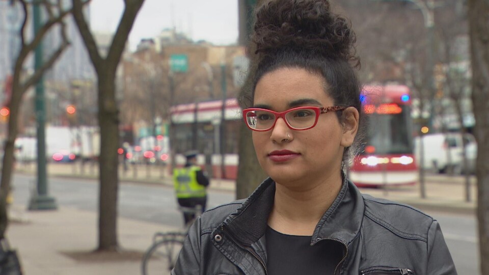 Une femme, portant des lunettes, à l'extérieur dans un quartier achalandé de Toronto. En arrière-plan, un tramway passe.