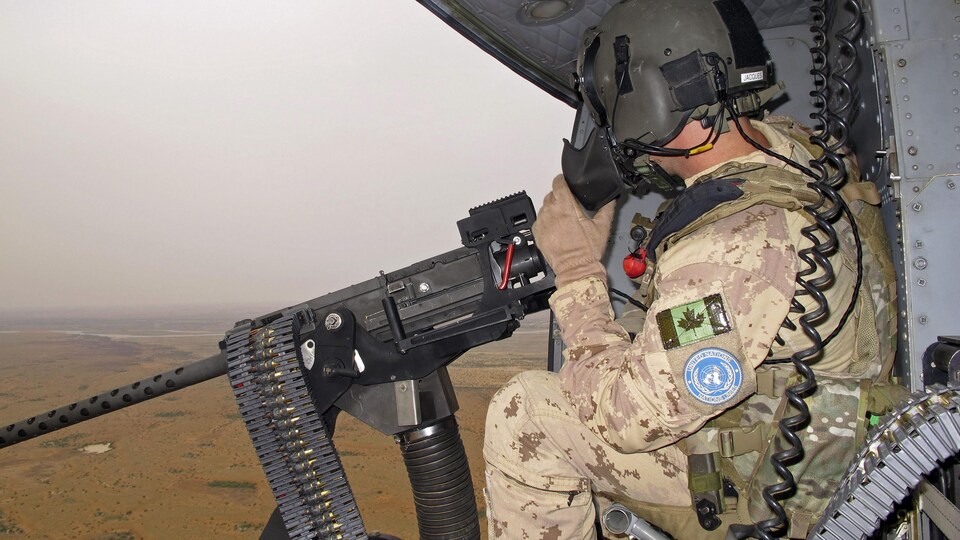 Un soldat canadien est assis dans un hélicoptère, devant un fusil mitrailleur.