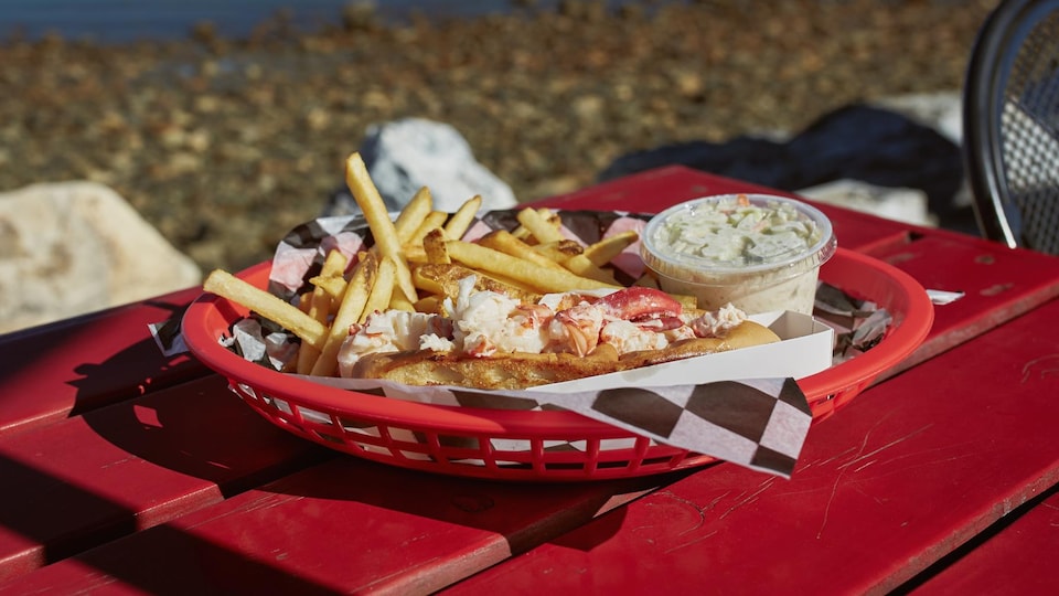 Un plat de guédille avec des frites et de la salade de chou est posé sur une table rouge devant des bateaux sur l'eau au loin.