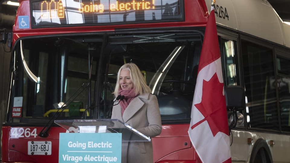 Catherine McKenna, in front of a bus and a Canadian flag, speaks in front of a lectern on which a sign reads: Electric turn.