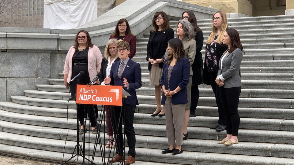 Dix femmes néodémocrates se tiennent sur les marches extérieures de l'Assemblée législative de l'Alberta, devant un podium.