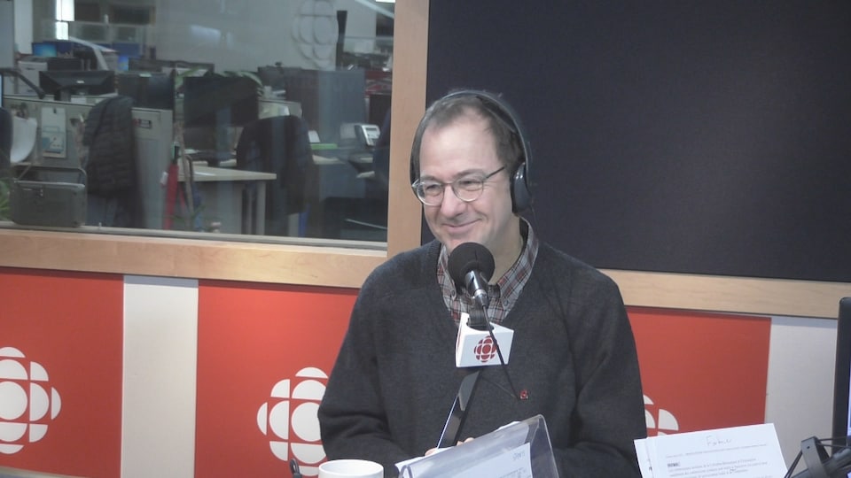Alain Fortier, un homme portant des lunettes, est assis dans un studio de radio, devant un microphone, avec des écouteurs sur la tête.