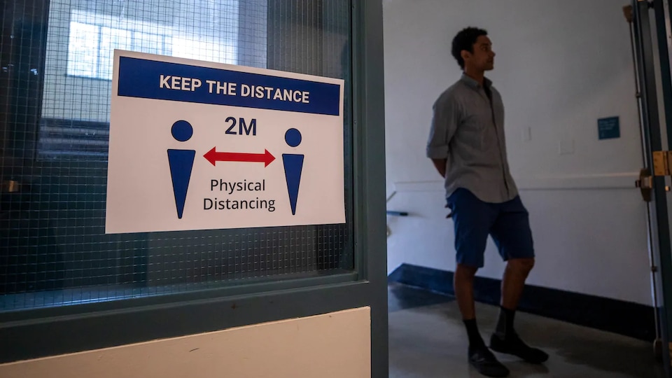 Une affiche placardée sur une fenêtre à côté de laquelle passe un jeune homme indique, en anglais, qu'il faut maintenir une distance physique d'au moins deux mètres entre les personnes.