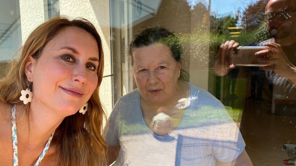 Deux femmes, dont l'une, âgée, se trouve derrière une vitre, posent pour une photo de famille.