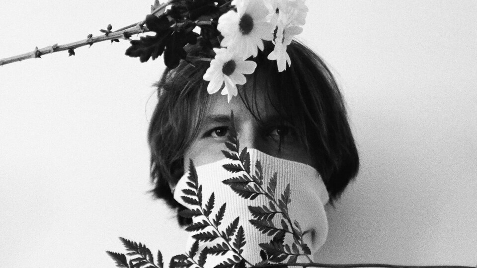 Le jeune homme porte un col roulé par-dessus son nez. Il a une branche de fougère devant le bas du visage et un bouquet de fleurs à la hauteur du front.