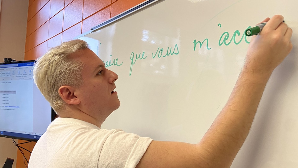 Le professeur Zacharie Bonneau écrit une phrase en français sur un tableau blanc d'école.