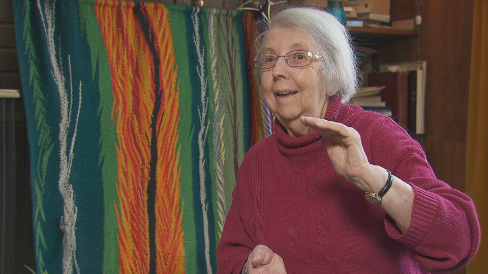 Une dame âgée assise dans une salle devant une ceinture fléchée dans l’arrière-plan.