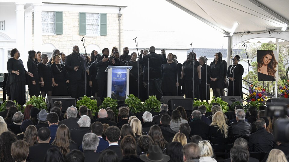 Un groupe de personnes habillées en noir chantent sur une scène surélevée. Devant elles, une foule, également vêtue de noir, les écoute.
