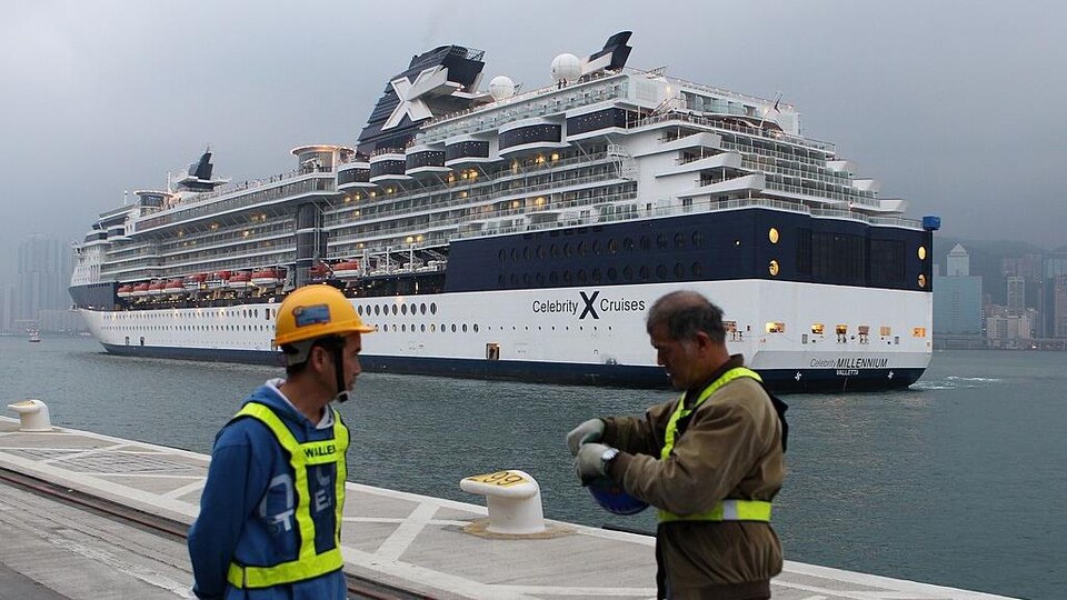 Deux travailleurs se tiennent sur le quai. Derrière eux, le bateau Celebrity Millennium se déplace tranquillement sur l'eau.