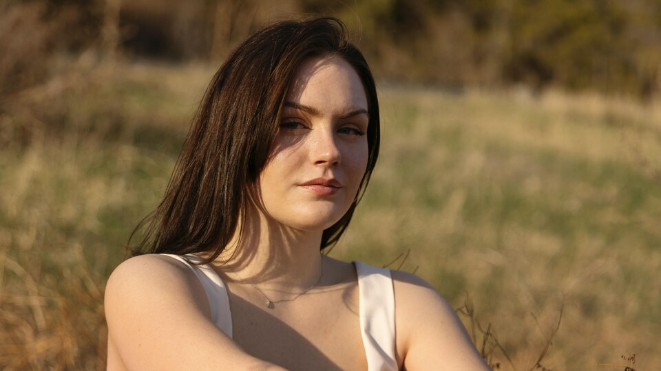 Céleste Beaupré, participante à Polyfonik 2023, est assise dans un champ et elle sourit à la caméra. 