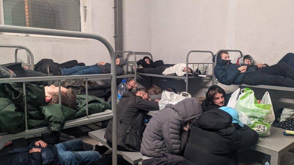 On voit des hommes entassés dans la cellule du centre de détention. Certains sont couchés sur des lits superposés, d'autres assoupis à une table.