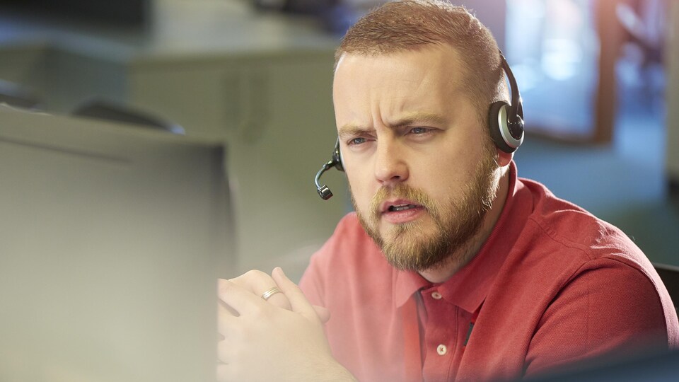 Un homme assis avec un casque d'écoute devant un ordinateur, dans un centre d'appels. Il a les sourcils froncés et écoute avec attention.