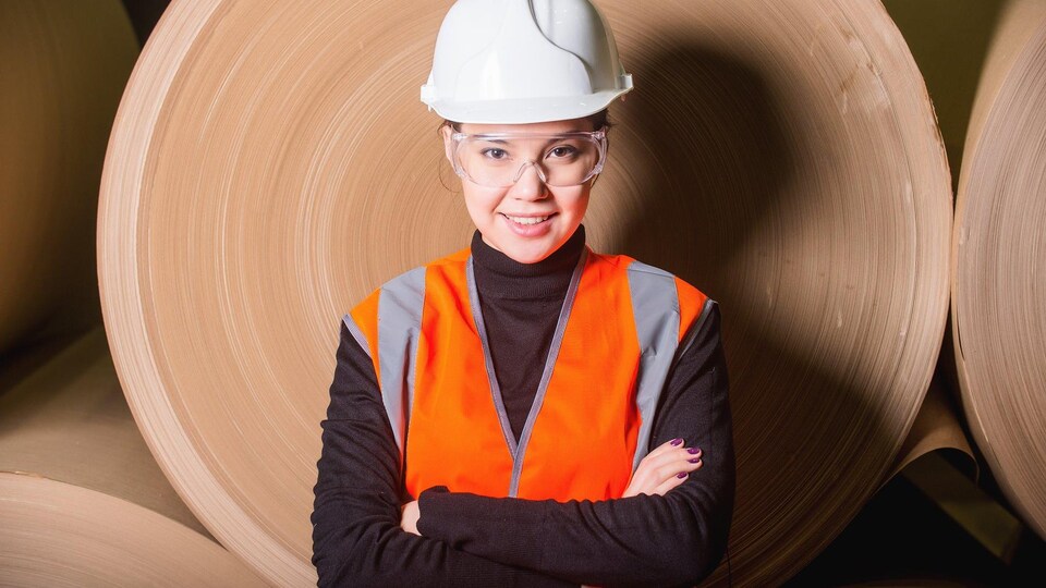 Une jeune femme sourit à la caméra, les bras croisés. Elle porte un dossard orange, des lunettes de sécurité et un casque blanc de protection. Derrière elle, d'immenses rouleaux de papier. Le Centre de formation professionnelle Pavillon-de-l'Avenir de Rivière-du-Loup offre entre autres le programme Pâtes et papiers – Opérations, plus de 500 heures en alternance travail-études.