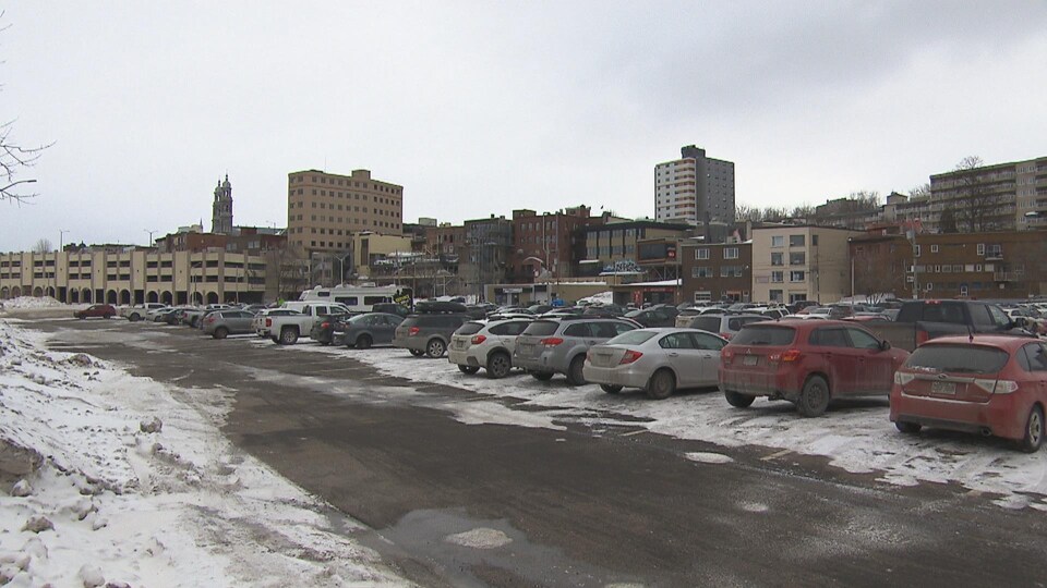Un stationnement avec des véhicules. Derrière, on peut voir le centre-ville de Chicoutimi.