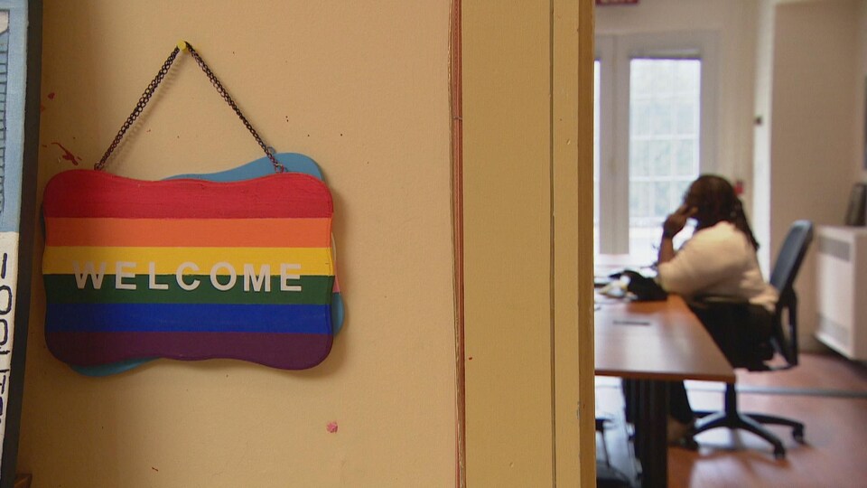 Au premier plan, une affiche aux couleurs de l'arc-en-ciel, posée sur le mur, indique "Welcome". À l'arrière-plan, une femme assise à une table parle au téléphone.