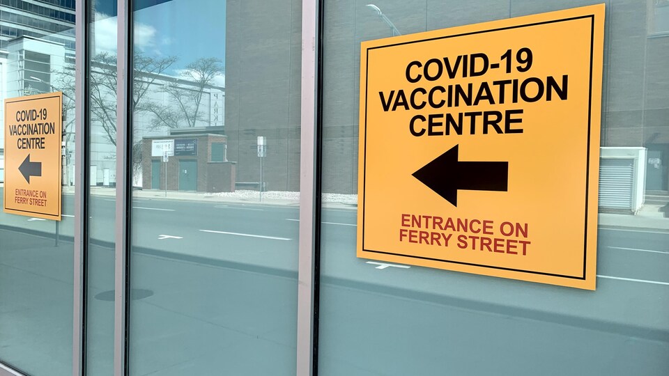 Des affiches indiquent la direction du centre de vaccination. Elles sont collées sur les fenêtres d'un bâtiment.