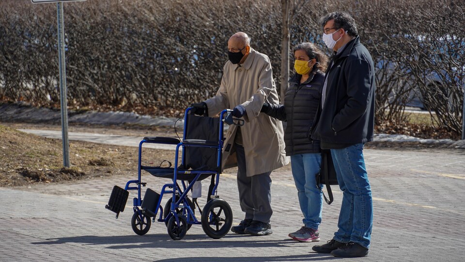 Des personnes accompagnent un parent qui marche avec une chaise roulante.