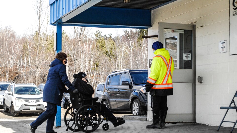 Une personne dans un fauteuil roulant est poussée vers un bâtiment. Un gardien de sécurité tient une porte.