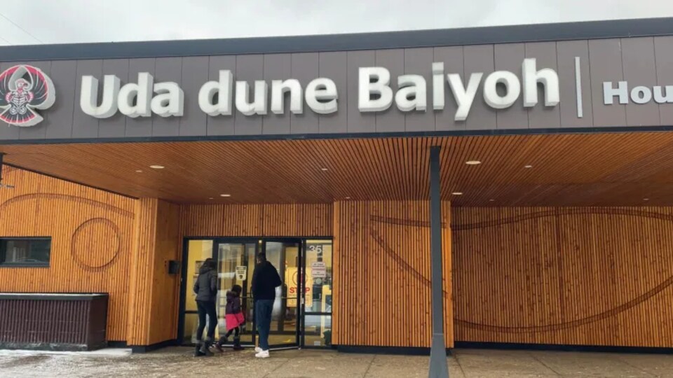 Deux adultes et un enfant entrent dans un bâtiment où il est inscrit Uda dune Baiyoh, (Maison des ancêtres, en français).