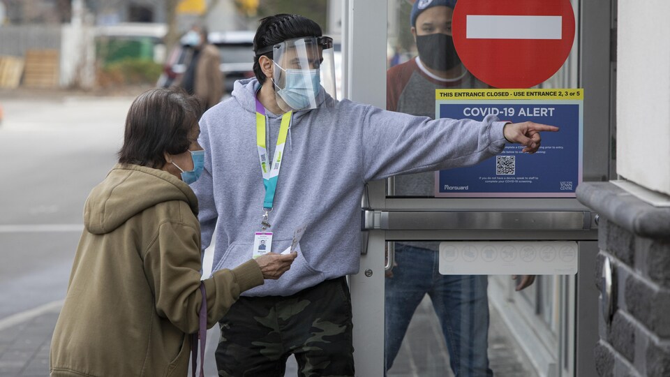 Un homme qui porte un masque et une visière indique à une femme âgée où elle doit aller pour se faire vacciner.