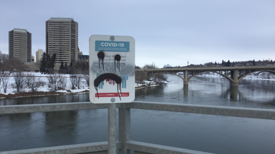 Un affiche au sujet de la COVID-19 barrée d'un graffiti représentant un visage triste avec, en arrière-plan, le centre-ville de Saskatoon, en Saskatchewan.