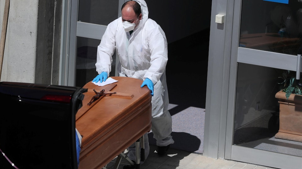 Un homme en tenue de plastique, masqué et muni de gants, pousse un cercueil dans un corbillard.
