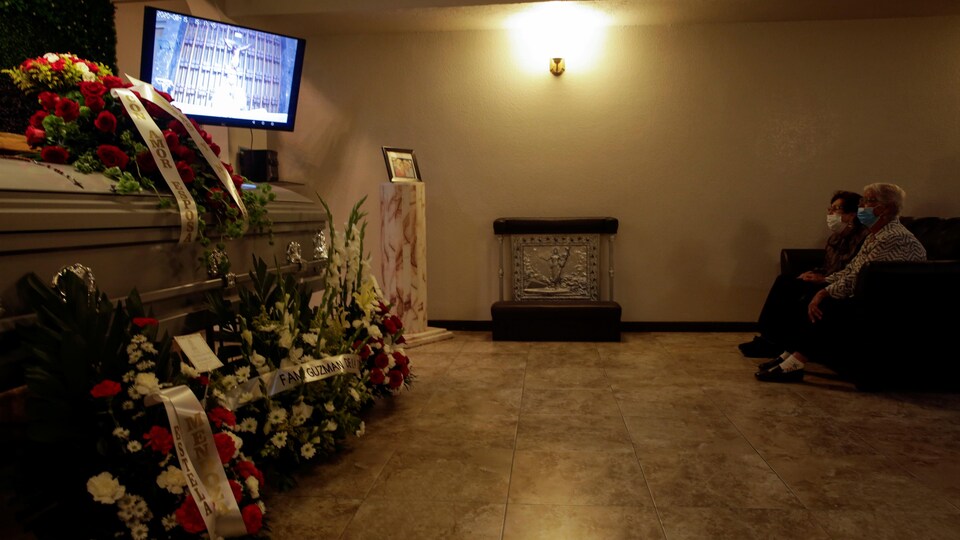 Deux femmes masquées sont assises devant un cercueil et une télévision. 