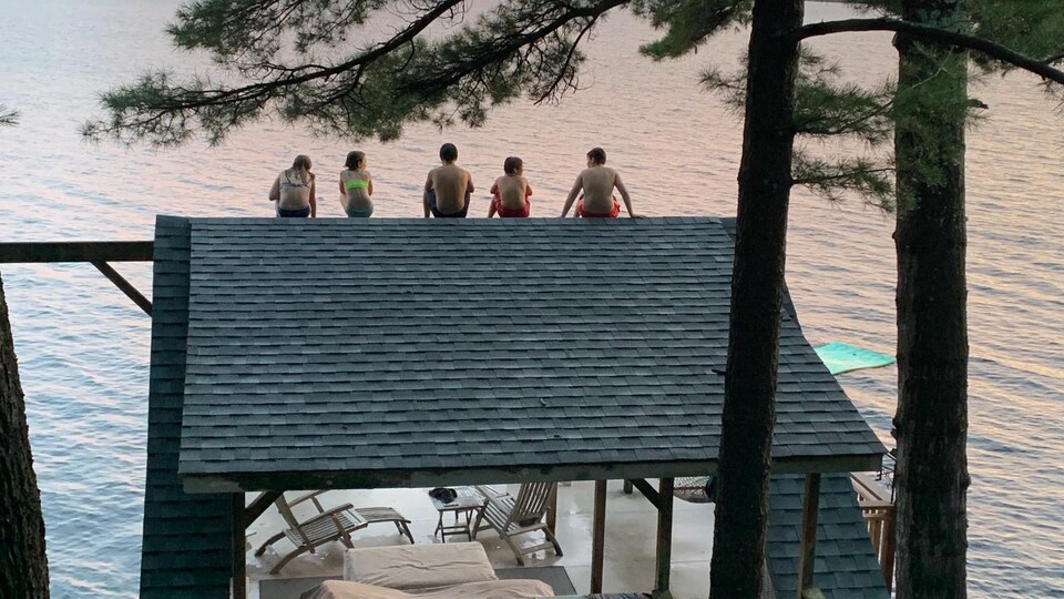 Cinq enfants sont sur le toit du cabanon d'un chalet devant un lac.