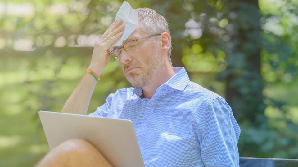 Man suffers from heat while working with laptop in the park and wiping his forehead with tissue
