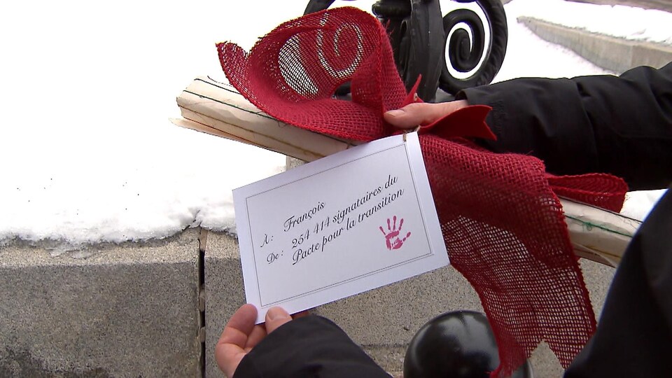 Des mains tiennent un rouleau de papier orné d'un ruban rouge et une carte disant « À François, de 254 414 signataires du Pacte pour la transition ».