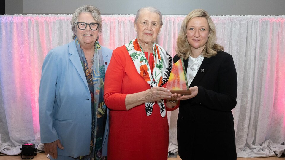Deux femmes ministres entourant la lauréate de 86 ans Christine Rioux-Ross. Cette dernière porte une robe et un long foulard fleuri noué. Elle sourit. 