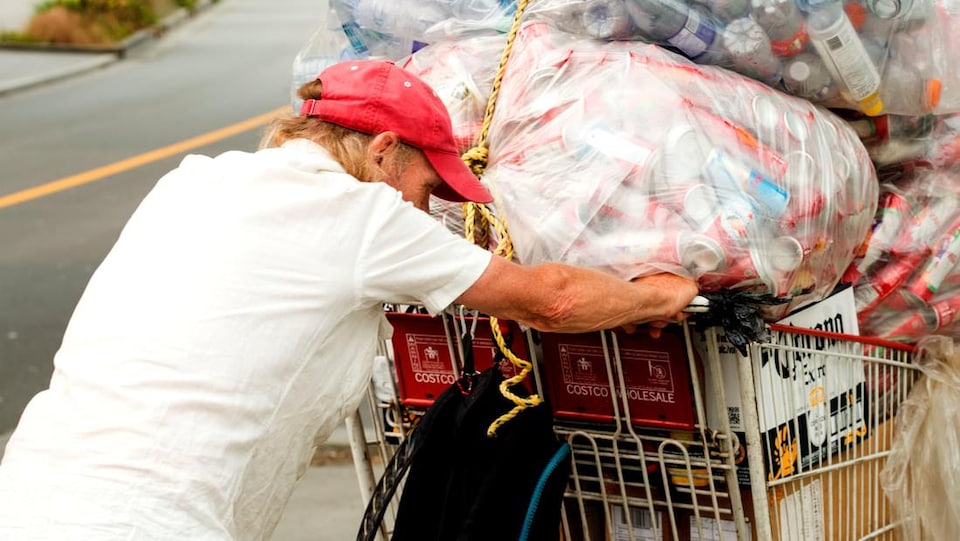 Un homme portant une casquette rouge pousse un chariot rempli de gros sacs de bouteilles et de cannettes qui sont destinés à être recyclés.