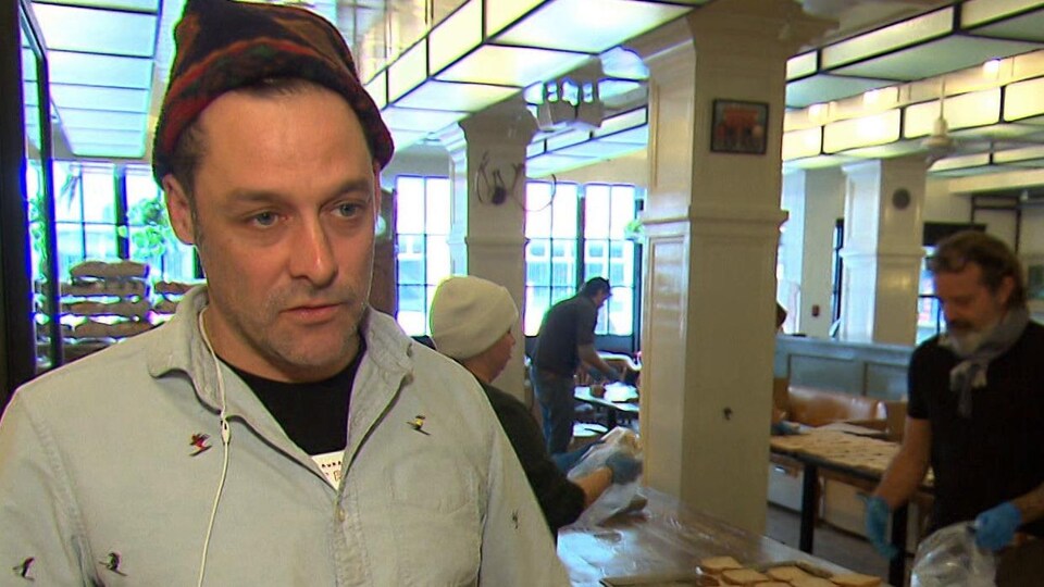 Le chef parlant à la caméra avec en arrière-plan, des bénévoles qui préparent des sandwichs.